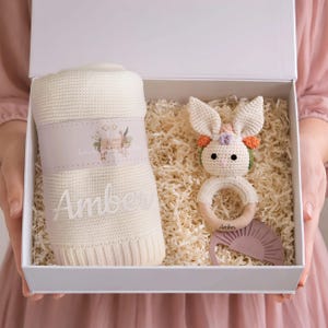 May include: A white gift box filled with shredded paper, holding a cream-colored baby blanket with the name "Amber", a crochet bunny rattle, and a pink teether. The blanket has a floral design.