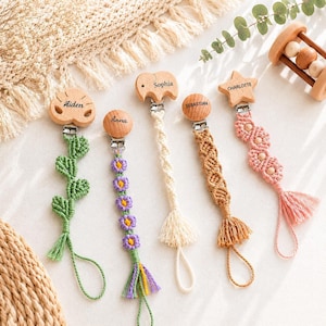 May include: Five macrame pacifier clips with wooden accents. Each clip features a unique shape: a leaf, a circle, an elephant, a circle, and a star, engraved with the names Aiden, Anna, Sophia, Sebastian, and Charlotte. The cords are crafted in green, purple, cream, tan, and pink.