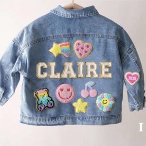 May include: A light blue denim jacket featuring the name CLAIRE in large white letters with gold trim. The back is decorated with colourful embroidered patches including a rainbow star, a pink heart, a teddy bear, a smiley face, cherries, and a flower.
