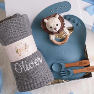 Könnte beinhalten: Ein Baby-Geschenkset mit einer grauen Strickdecke mit dem Namen "Oliver", einem blauen Silikon-Lätzchen, einem gehäkelten Löwenrassel und einem Löffel- und Gabel-Set. Das Set wird in einer weißen Box präsentiert.