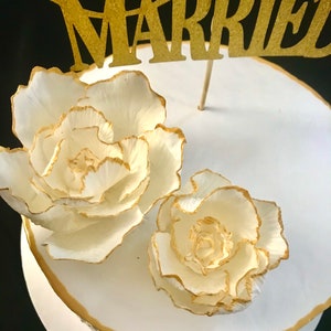 May include: A white cake with gold accents and two white sugar flowers. The cake is topped with a gold glitter topper that says "Married".