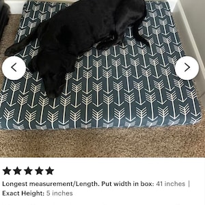 May include: A dark gray dog bed with a white arrow pattern. The bed is 41 inches wide and 5 inches high. A black dog is lying on the bed.