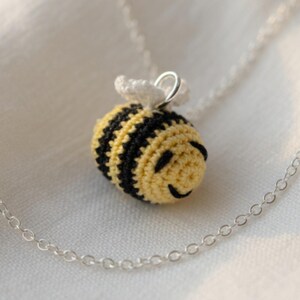 May include: A crocheted bee pendant necklace with a silver chain. The bee is yellow and black with a smiling face and white wings. The necklace is displayed on a white surface.