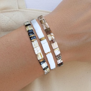 May include: Three beaded bracelets in neutral tones are stacked on a wrist. The bracelets feature white, black, and brown square beads. The background includes a white top and a beige tote bag.