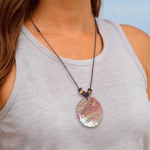 May include: An oval pendant necklace with an iridescent, shimmering surface. The pendant hangs from a black cord with wooden beads. The person is wearing a grey vest top and blue jeans, with a beach and ocean in the background.