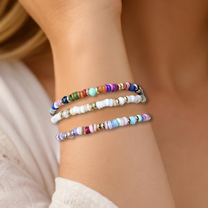 May include: Three beaded bracelets in various colors, including white, blue, purple, and gold. The bracelets are stacked on a wrist, showcasing a mix of colorful beads. The bracelets are a fashion accessory.
