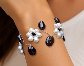 May include: A floral bracelet featuring white pearl-like beads and black onyx-like stones. The bracelet has a delicate, wire-like design with flower-shaped clusters. The bracelet is worn on a wrist, showcasing its elegant design.