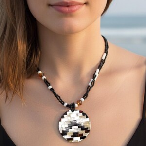 May include: A black beaded necklace with a large, circular pendant. The pendant has a mosaic design in black, white, and tan. The necklace is worn with a black top.