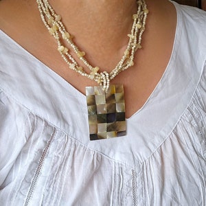 May include: A multi-strand necklace made of white beads with a large square pendant made of iridescent mother of pearl tiles.