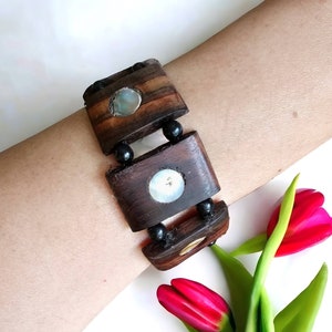 May include: A wooden bracelet with three round, iridescent shell inlays. The bracelet is made of dark brown wood and has black beads between each section.