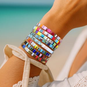 May include: A wrist adorned with multiple bracelets, each composed of small, colourful, square beads. The bracelets display a spectrum of hues, including red, pink, yellow, blue, and green. The backdrop reveals a beach with turquoise water.