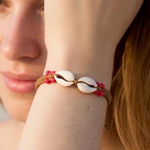May include: A woman wearing a seashell bracelet. The bracelet has two white seashells and red beads on a brown cord. The background shows a beach and the sea. The bracelet is a piece of jewellery.