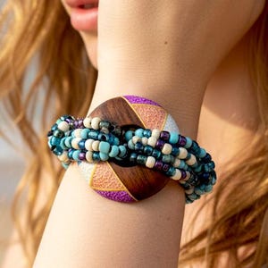 May include: A bracelet featuring a circular dark wood centerpiece with purple and gold textured accents. The wood piece is connected to multiple strands of small glass beads in shades of turquoise, navy blue, white, and black, wrapped around the wrist.