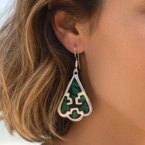 Könnte beinhalten: Silberne Ohrhänger mit dreieckiger Form. Die Ohrringe haben eine dunkelgrüne Einlage mit geometrischem Design. Sie hängen an einem silbernen Haken. Der Hintergrund zeigt eine Strandszene.