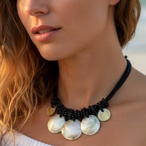 May include: A black beaded necklace featuring large, round, iridescent shell pendants and small gold-coloured discs. The necklace is worn with a white off-the-shoulder top. The background is a blurred beach scene.