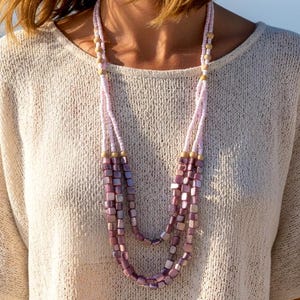 May include: A layered necklace with light purple square beads and gold accents. The necklace is worn over a cream-colored, short-sleeved top. The background features a beach and ocean.