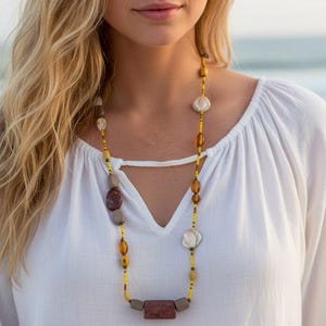 May include: A long beaded necklace with a variety of stone and shell beads in shades of brown, yellow, and white. The necklace is worn over a white blouse with a V-neck. The background is a beach scene with the ocean.