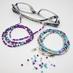 May include: A pair of spectacles with a silver frame sits above two beaded eyeglass chains. One chain is purple, blue, and white, and the other is blue, orange, and white. A few loose beads are scattered below.