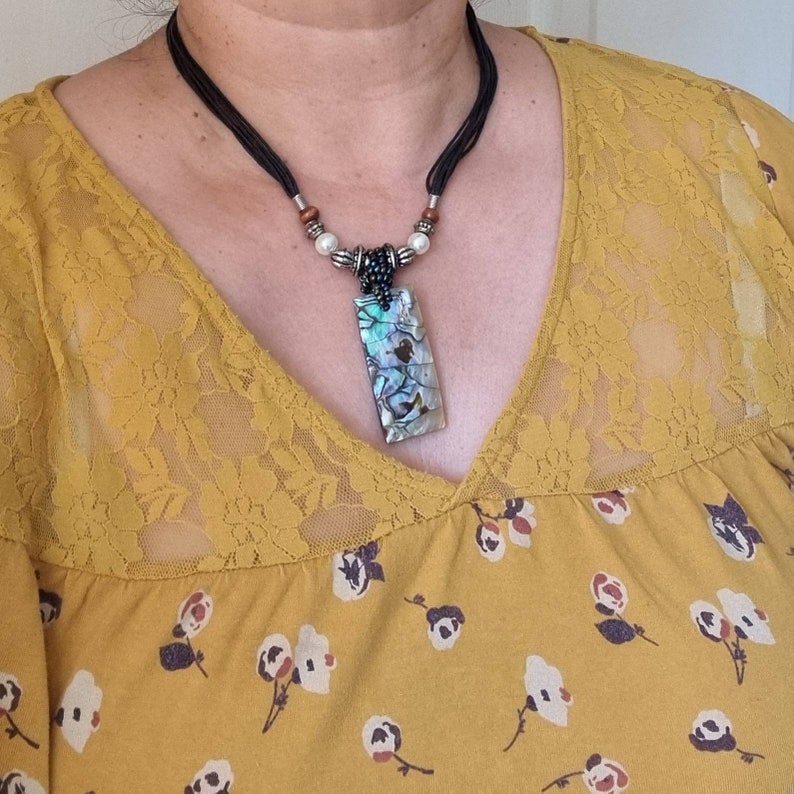 May include: Abalone shell pendant necklace. The rectangular pendant features iridescent blues, greens, and purples. The necklace has a black cord with decorative beads and a clasp. The necklace is worn over a yellow floral top.