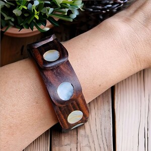 May include: A dark brown wooden bracelet with three oval-shaped mother-of-pearl inlays and two circular inlays. The bracelet is worn on a wrist, showcasing the natural wood grain and the iridescent quality of the inlays.