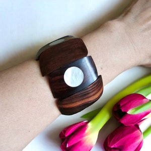 May include: A wooden bracelet with a dark brown wood finish and a white mother-of-pearl inlay. The bracelet is made of multiple pieces of wood that are joined together. The bracelet is shown on a person's wrist.