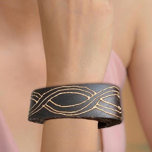 May include: A black leather bracelet with a gold-coloured geometric design. The bracelet is worn on a person's wrist.