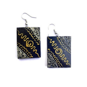 May include: A pair of black wooden earrings with gold painted designs. The earrings have a rectangular shape and feature a swirl pattern and wavy lines.