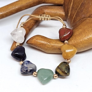 May include: A bracelet with heart-shaped stones in various colours, including clear, red, yellow, green, blue, and tiger's eye. The stones are strung on a gold-coloured cord with small gold beads. The bracelet is displayed on a wooden surface.