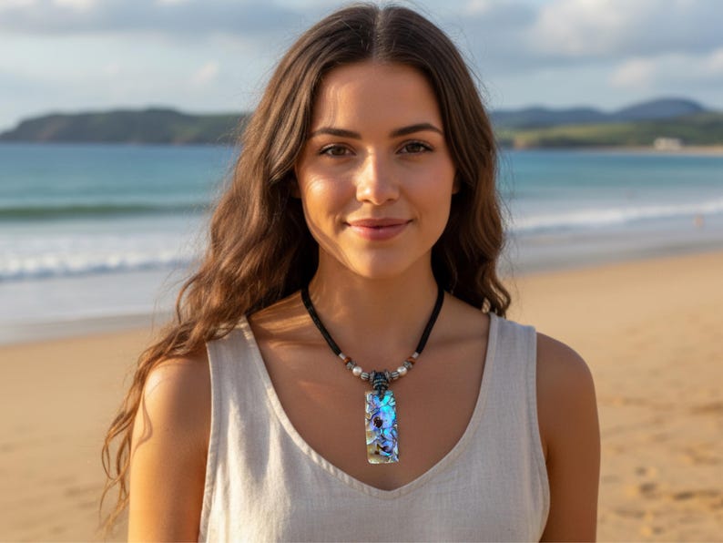May include: A rectangular pendant necklace with a black cord. The pendant has a colorful, iridescent design. The necklace is worn against a light-colored top, with a beach and ocean in the background.