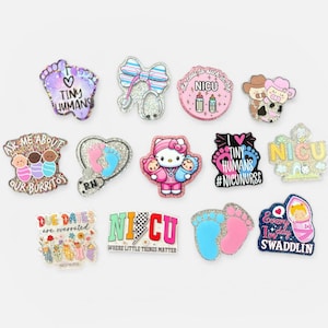 Puede incluir: Una colección de coloridos y brillantes dijes con varios diseños y texto. Los diseños incluyen pies de bebé, lazos, corazones y los textos "Tiny Humans", "NICU" y "Due Dates are overrated."