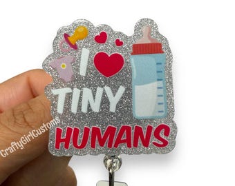 May include: A glittery badge reel with the phrase "I LOVE TINY HUMANS" in white and red. The design includes a pacifier, hearts, a baby bottle, and a onesie. The reel is silver and has a clear plastic clip.