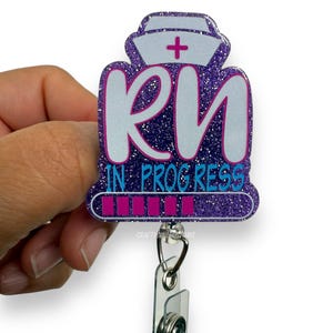 Puede incluir: Un carrete de insignia RN con purpurina morada con un diseño de gorro de enfermera. La insignia presenta las letras "RN" en blanco con un contorno rosa y las palabras "IN PROGRESS" en azul. Una barra de progreso rosa en la parte inferior.