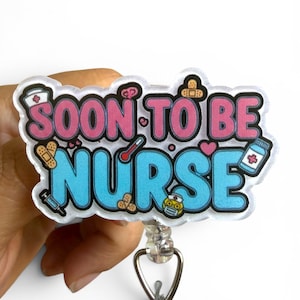 May include: A badge reel with the words "SOON TO BE NURSE" in pink and blue, with medical-themed illustrations. The badge reel has a clear, plastic covering and a silver clip.