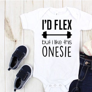 May include: White baby onesie with black text that reads "I'd flex but I like this onesie" with a black dumbbell graphic above the text. The onesie is on a wooden surface with a pair of blue baby shoes, blue jeans, and a baseball.