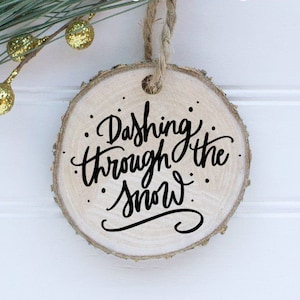 May include: A round wooden ornament with a white background and black calligraphy that reads "Dashing Through The Snow".
