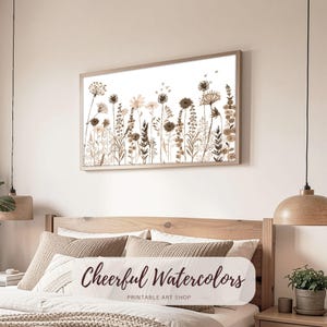 Puede incluir: Una impresión enmarcada de acuarela que representa un campo de flores silvestres en tonos marrones y beige sobre un fondo blanco. La obra de arte se exhibe sobre una cama. El texto "Cheerful Watercolors" es visible en una almohada.