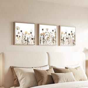 May include: A set of three square wall art prints featuring botanical illustrations of wildflowers and grasses in shades of beige, brown, and cream. Each print is encased in a light-coloured wooden frame, displaying detailed floral silhouettes.
