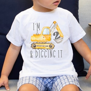 May include: A white t-shirt featuring a yellow excavator design and the text "I'm 2 & digging it".