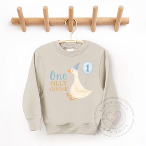 May include: A pale beige sweatshirt featuring a cartoon goose wearing a party hat and holding a blue balloon with the number 1. The text "One Silly Goose" is above the goose.