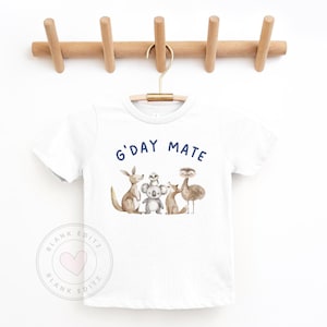 May include: White t-shirt with the text "G'DAY MATE" in navy blue. The shirt features a watercolor illustration of Australian animals: a kangaroo, koala, frog, fox, and emu. The shirt is hanging on a wooden coat rack.