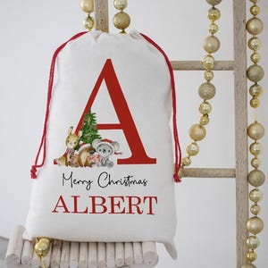 Puede incluir: Bolsa de regalo navideña blanca con cordón rojo, con una gran letra "A" roja y el nombre "ALBERT" en rojo. La bolsa está decorada con un árbol de Navidad y animales de dibujos animados. El texto "Feliz Navidad" también está impreso en la bolsa.