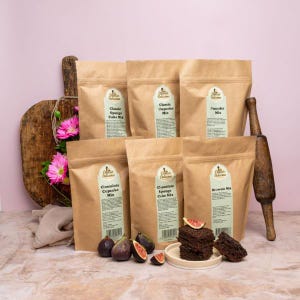 May include: Six brown paper bags with different cake mix labels. The labels have the text "Classic Sponge Cake Mix", "Classic Cupcake Mix", "Pancake Mix", "Chocolate Cupcake Mix", "Chocolate Sponge Cake Mix", and "Brownie Mix". There are also some figs and a plate of brownies in the image.