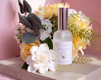 May include: A frosted glass spray bottle labeled Blue Gardens Beauty Gardenia Room and Linen Spray with a metallic cap, positioned next to a white gardenia flower and a bouquet of yellow roses and eucalyptus leaves.