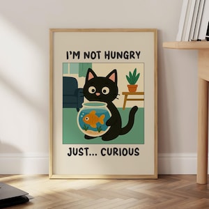 May include: A framed art print featuring a cartoon black cat holding a fishbowl with a goldfish. The print has the text "I'M NOT HUNGRY" above the cat and "JUST... CURIOUS" below. The background is a light green with a blue couch and a small table.