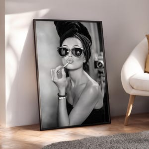 Op de afbeelding: Zwart-wit ingelijste print van een vrouw die lippenstift aanbrengt. Ze draagt een zonnebril en een zwarte handdoek op haar hoofd. De afbeelding leunt tegen een muur, met een witte stoel en een grijs tapijt op de achtergrond.