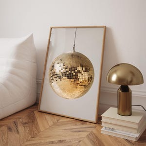 May include: A gold disco ball hanging from the ceiling in a framed print. The print is on a white background and is sitting on a wooden floor.