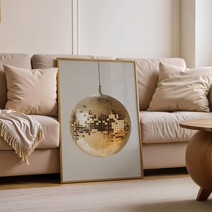 May include: A gold disco ball hanging from the ceiling in a framed print. The print is hanging on a wall above a beige sofa with pillows.