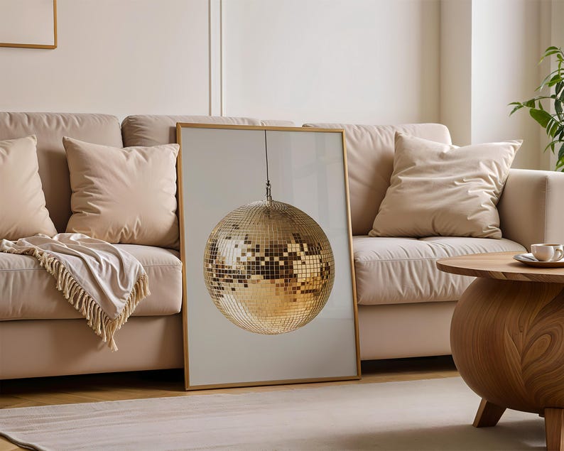 May include: A gold disco ball hanging from the ceiling in a framed print. The print is hanging on a wall above a beige sofa with pillows.