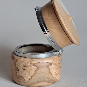 May include: A wooden box with a hinged lid and a silver metal trim. The box is made of light brown wood with a natural wood grain pattern. The lid is open and the inside of the box is visible.