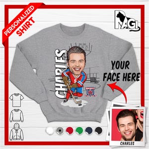 May include: Personalized gray sweatshirt with a cartoon illustration of a hockey player. The player is wearing a red and blue jersey with the number 5. The text "Charles" is written vertically on the left side of the image. The text "Your Face Here" is written on the right side of the image with an arrow pointing to the player's face.